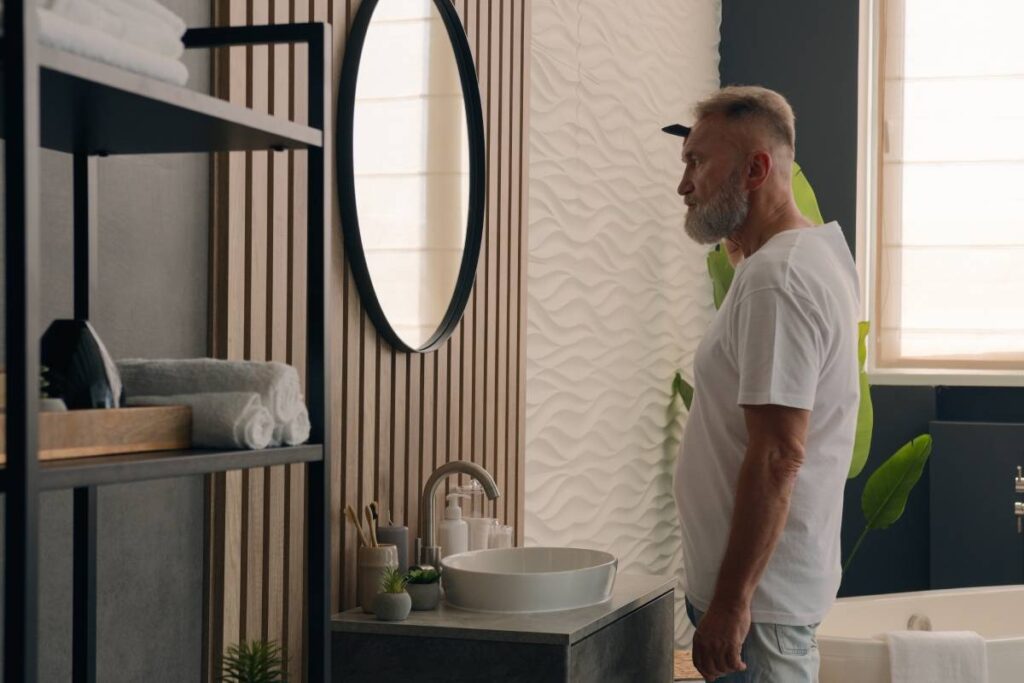 Caucasian bearded old senior man mature middle aged elderly retired male looking at mirror in bathroom combing gray hair with hairbrush comb beard alopecia healthy hairstyle morning beauty routine.